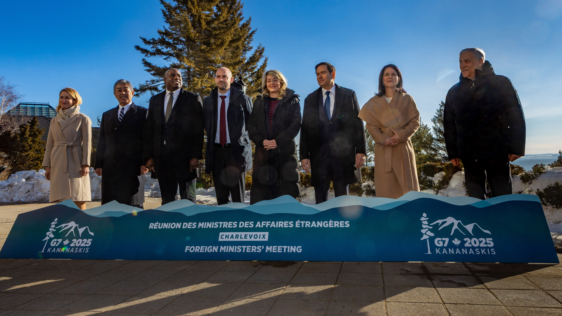 The eight ministers are bundled up against the cold with a sign for their meeting in front of them and a bright blue sky behind them.