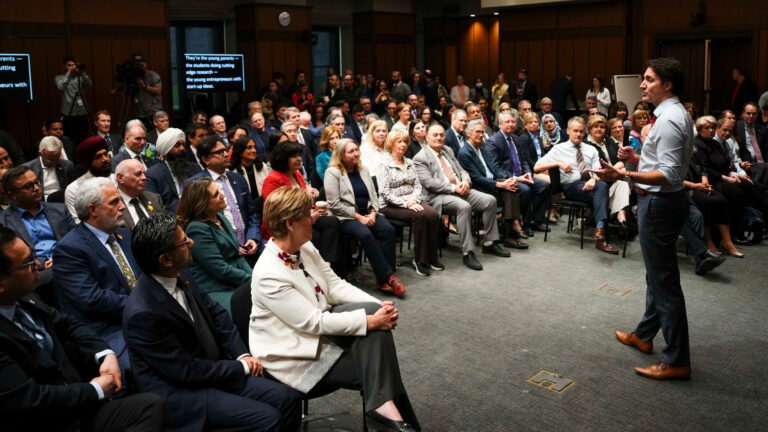 Trudeau in shirtsleeves stands and speaks in front of MPs gathered in a conference room. Teleprompters in the back of the room show Trudeau’s speech.