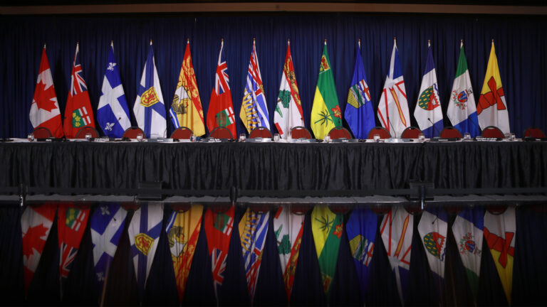 The flags of the provinces and territories are lined up on poles in front of a blue curtain in a conference room.