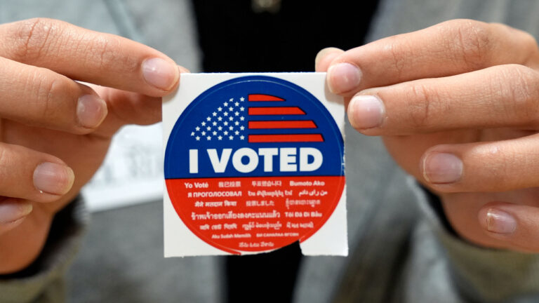 un homme tient un autocollant aux couleurs du drapeau américain sur lequel on peut lire la mention « J’ai voté » dans plusieurs langues.