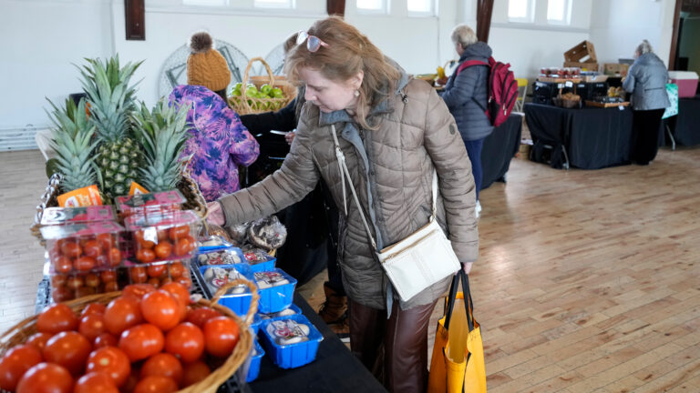 Une femme en manteau beige se penche pour prendre des fruits et légumes sur un étalage.