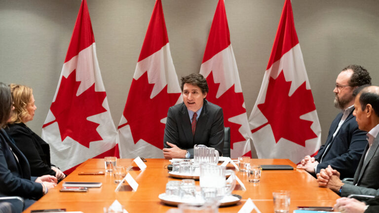 Justin Trudeau sits at the head of a boardroom talking to men and women in business attire. He has four Canadian flags behind him.