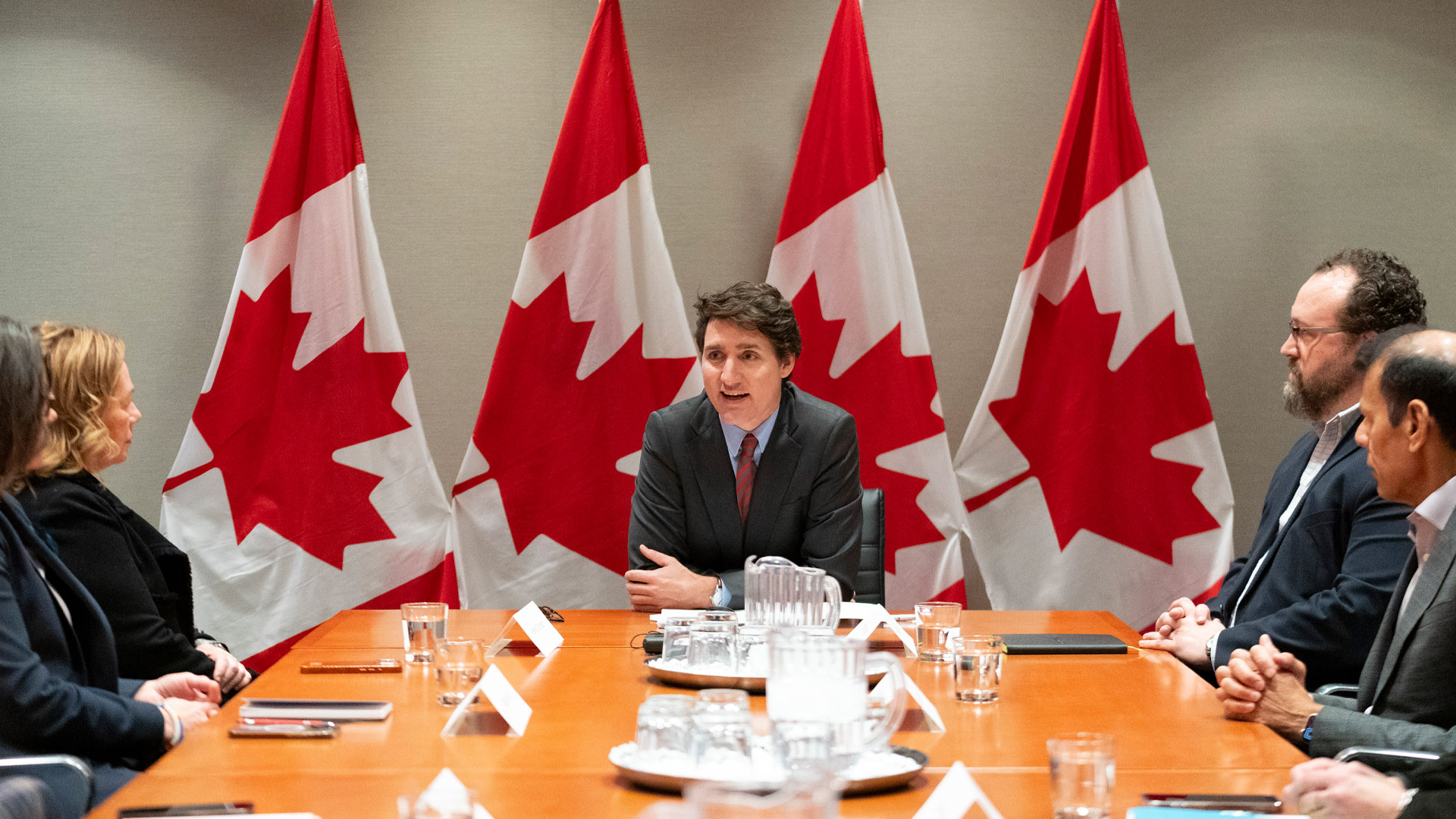 Justin Trudeau sits at the head of a boardroom talking to men and women in business attire. He has four Canadian flags behind him.