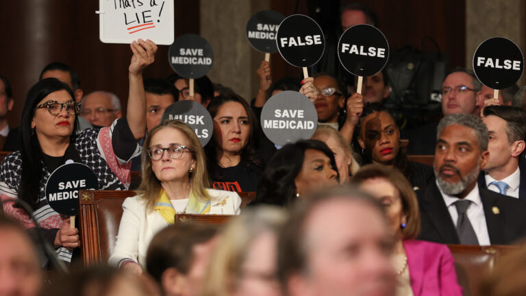Des membres du Parti démocrate, assis dans la salle du Congrès américain, tiennent des pancartes noires avec les messages écrits en blanc «SAVE MEDICAID», «FALSE» et «MUSK KILLS». Une élue au premier plan brandit un tableau blanc sur lequel est écrit «That's a LIE!» en lettres rouges et noires. Les visages des élus sont sérieux ou réprobateurs, illustrant une protestation silencieuse pendant une allocution présidentielle.