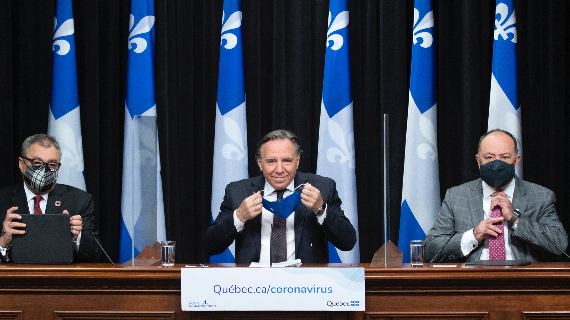 Three men wearing suits, seated at a desk in front of a row of Quebec flags, are about to give a press conference. They are wearing face coverings. The man in the middle removes his mask.