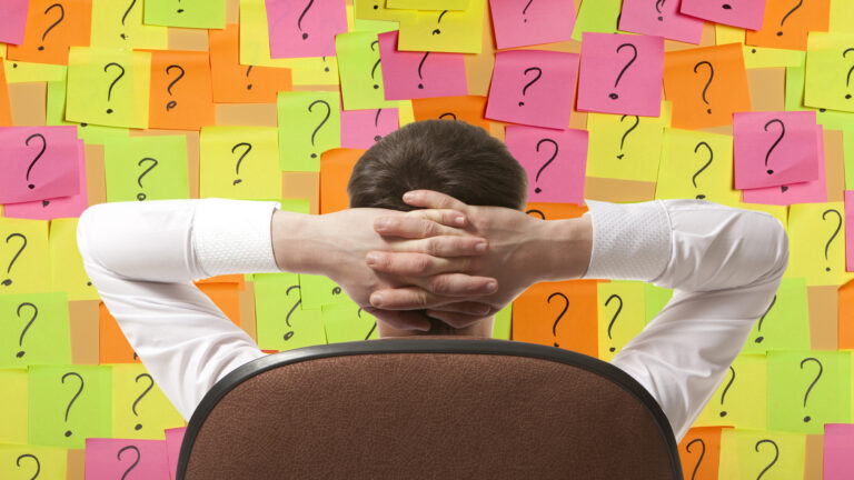 A photo illustration showing a man in a suite before a wall of sticky notes with question marks.