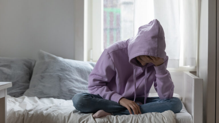 An adolescent girl sits cross-legged on a sofa next to a window with sheer white curtains. She’s downcast, leaning forward with one hand on her forehead and her elbow on her leg. She’s wearing jeans and a pastel-purple hoodie that covers her head.