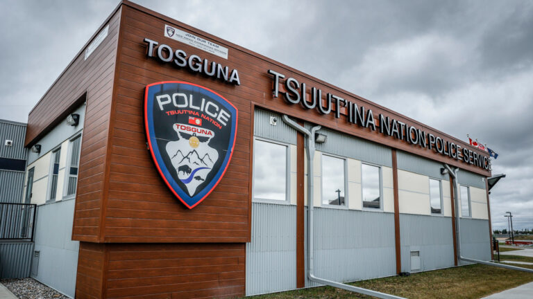 Modern Tsuut'ina Nation Police Service building with a wooden sign reading "TOSGUNA TSUUT'INA NATION POLICE SERVICE." A large police crest is displayed on the left, depicting mountains, a bison, and cultural symbols. Canadian and Indigenous flags fly from the roof under a cloudy sky.