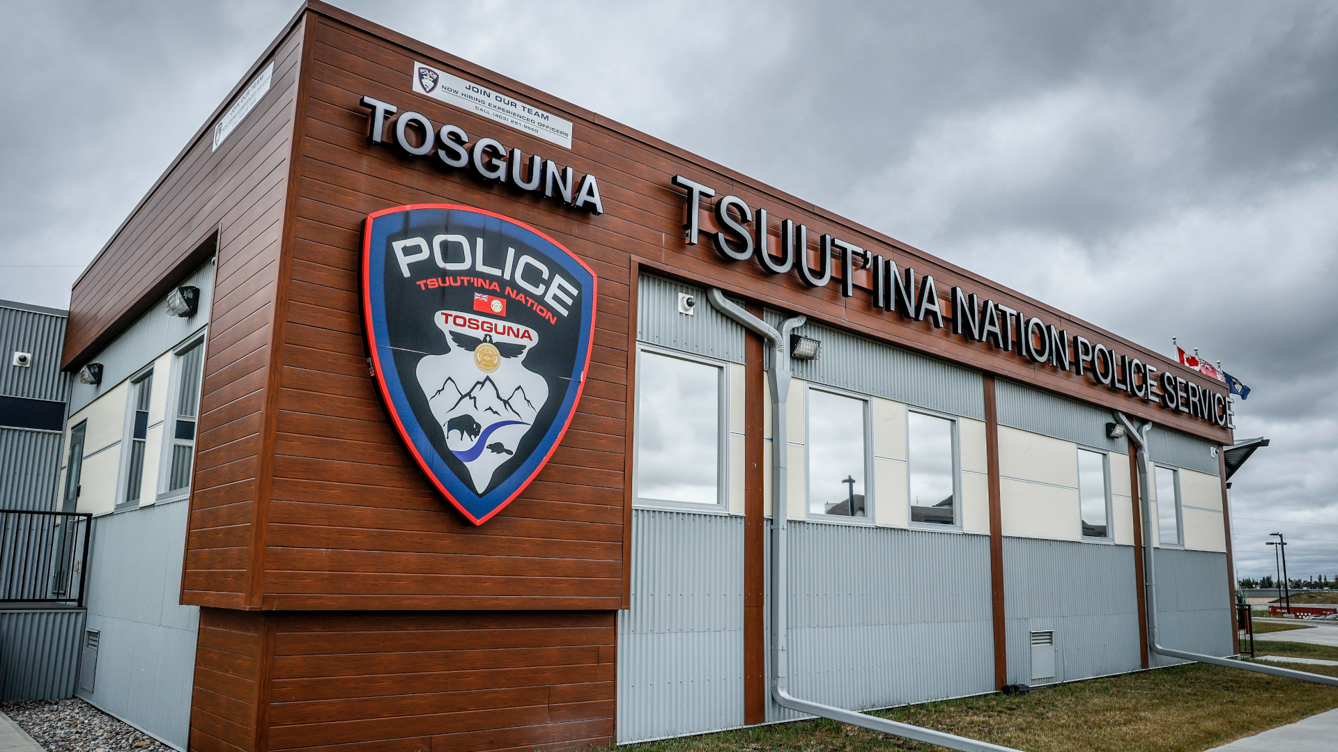 Modern Tsuut'ina Nation Police Service building with a wooden sign reading "TOSGUNA TSUUT'INA NATION POLICE SERVICE." A large police crest is displayed on the left, depicting mountains, a bison, and cultural symbols. Canadian and Indigenous flags fly from the roof under a cloudy sky.