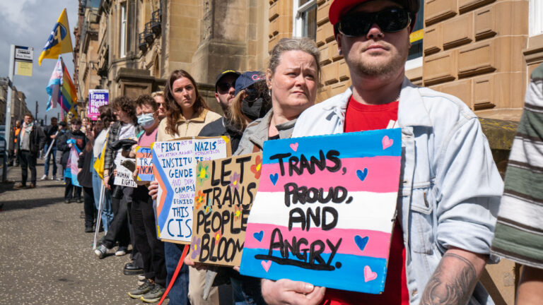 Manifestation pour les droits des personnes transgenres devant un bâtiment. Des manifestants tiennent des pancartes aux messages pro-trans comme « Trans, proud, and angry ». Drapeaux trans et LGBTQ+, ambiance pacifique, chien au premier plan.