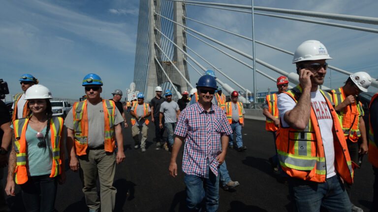 A dozen smiling people wearing orange safety vests and hard hats walk along the new span towards the camera.