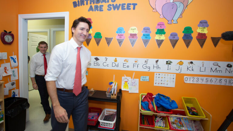 Trudeau enters a room in a daycare, with Furey behind him, passing through the doorway. The walls are bright orange and covered with stickers depicting pastel-coloured balloons and ice-cream cones with the dates of birthdays of the children written on the scoops of ice cream. Letters on the wall spell out “birthdays are sweet.” Other papers taped to the wall show the alphabet and numbers. Low shelves hold markers, crayons and other drawing supplies. The two men are smiling, and both wear dark pants, a white shirt and red ties.