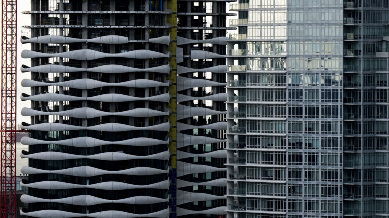 We see about 20 storeys of two towers, one complete with floor-to-ceiling glass, the other partially finished, with the five upper floors less complete. The balconies on the new building have a decorative material, curved sheets of light grey metal. Taken together, they resemble a pattern of undulating small ocean waves.