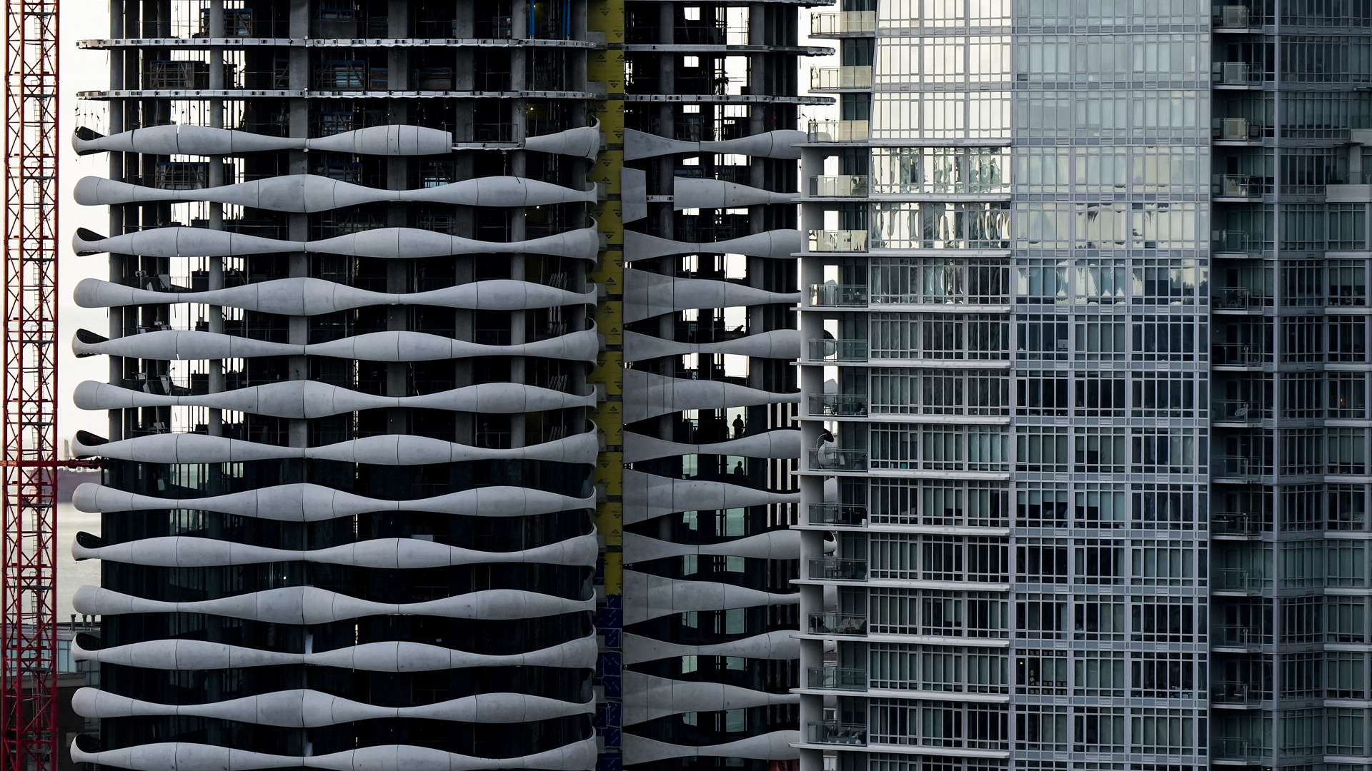 We see about 20 storeys of two towers, one complete with floor-to-ceiling glass, the other partially finished, with the five upper floors less complete. The balconies on the new building have a decorative material, curved sheets of light grey metal. Taken together, they resemble a pattern of undulating small ocean waves.