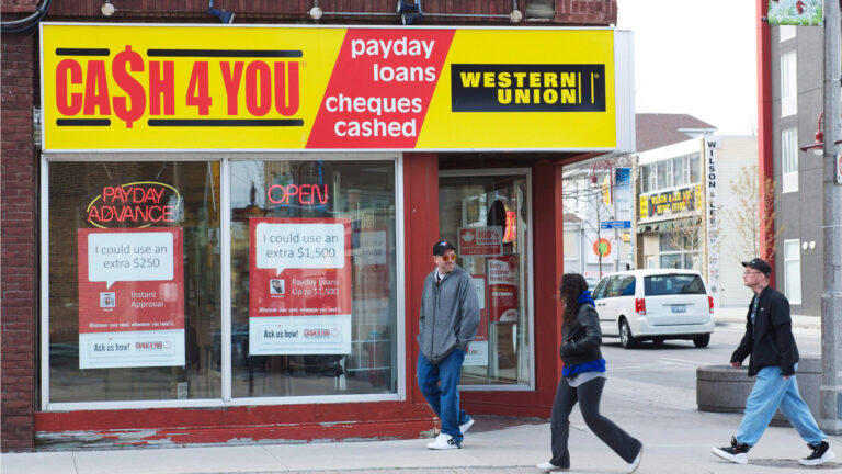 Three people are walking past an outlet with CA$SH4YOU and a Western Union logo on the front, with a “payday advance” neon sign in the window.