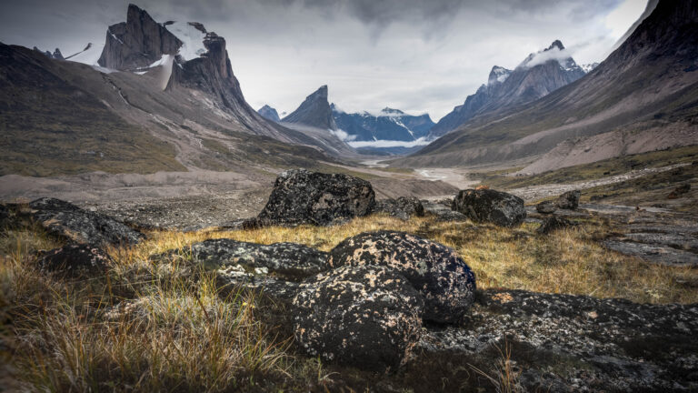 Dark mist swirls on a white sky, with dark mountains all around. Dark lichen-covered boulders sit amid golden tall grass in the foreground.