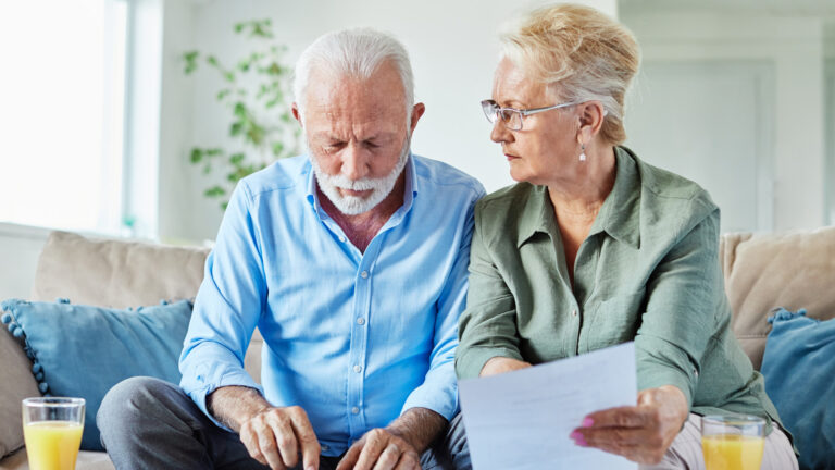 Un homme et une femme blancs d’un certain âge sont assis sur un canapé et consultent des feuilles de papier posées sur une table basse devant eux. Ils utilisent une calculatrice