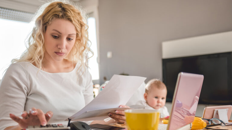 A woman works a calculator at a table with a laptop computer while a baby watches a screen in the background.
