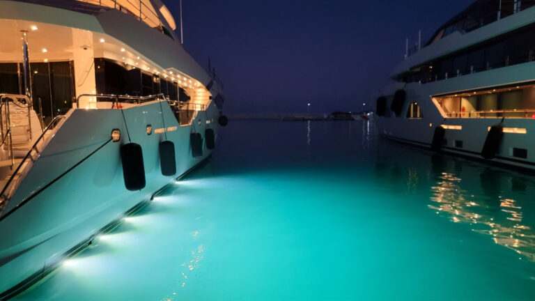 Two yachts are berthed at a marina at night. The water between the two hulls is lit up by lights from the waterline of the boat on the left, giving it a bright turquoise colour. Both boats are white fibreglass with several levels of decks. Pot lights on each boat cast a warm glow.