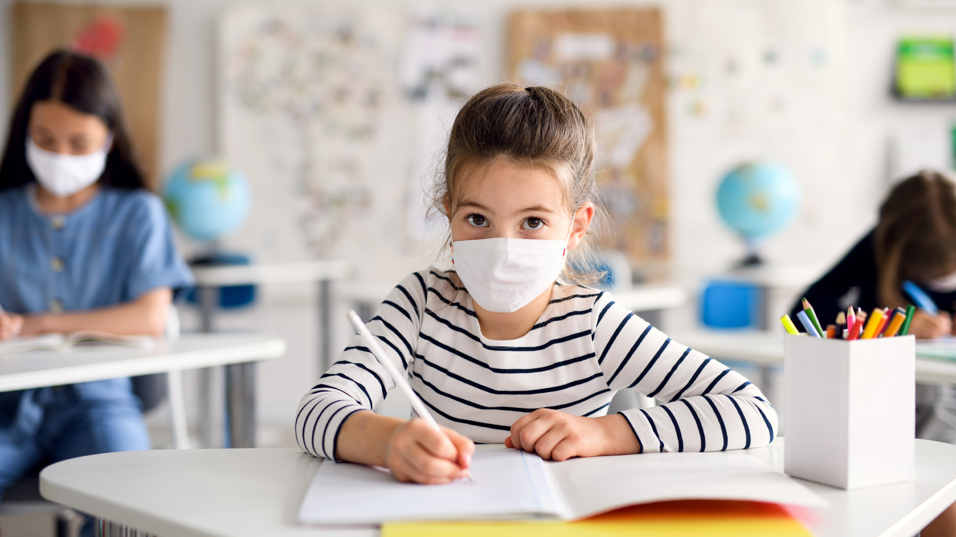 A young girl in a classroom wears a mask. She’s doing schoolwork at a desk, with two other students at desks nearby.
