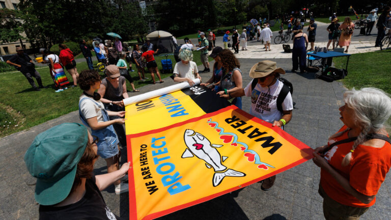 Un petit groupe de manifestants, dont une femme en fauteuil roulant battant un petit tambour, se rassemble à côté d'une banderole représentant un poisson et des cœurs avec les mots « Nous sommes ici pour protéger l'eau ».