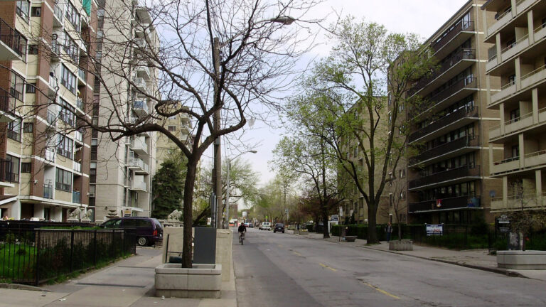 Looking down a street lined with large apartment buildings on both sides. They are about seven storeys high, built of brick with concrete balconies. The street is quiet. No passing cars, no parked cars, no pedestrians. A cyclist is approaching. Several trees are growing from a series of concrete planters.