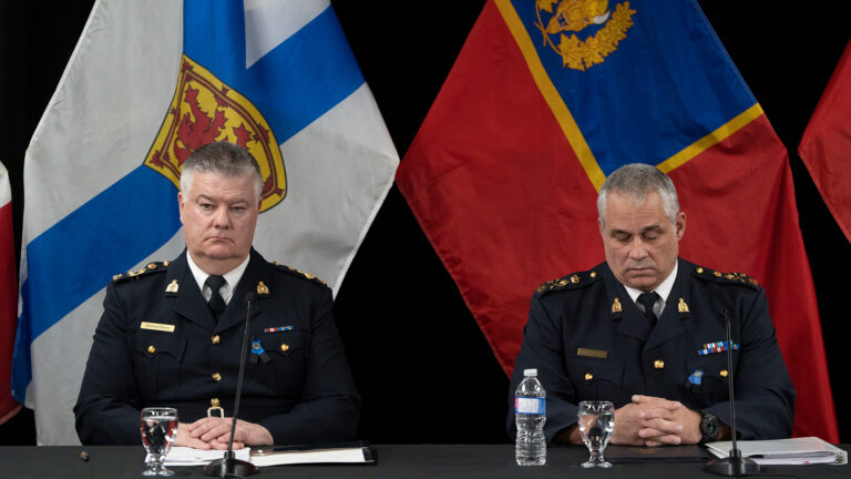 Both men sit at a long table covered in a black tablecloth. They are both wearing black uniforms with pins signifying various honours and achievements. Behind them are two large flags, one the Nova Scotia flag, the other the flag of the RCMP.