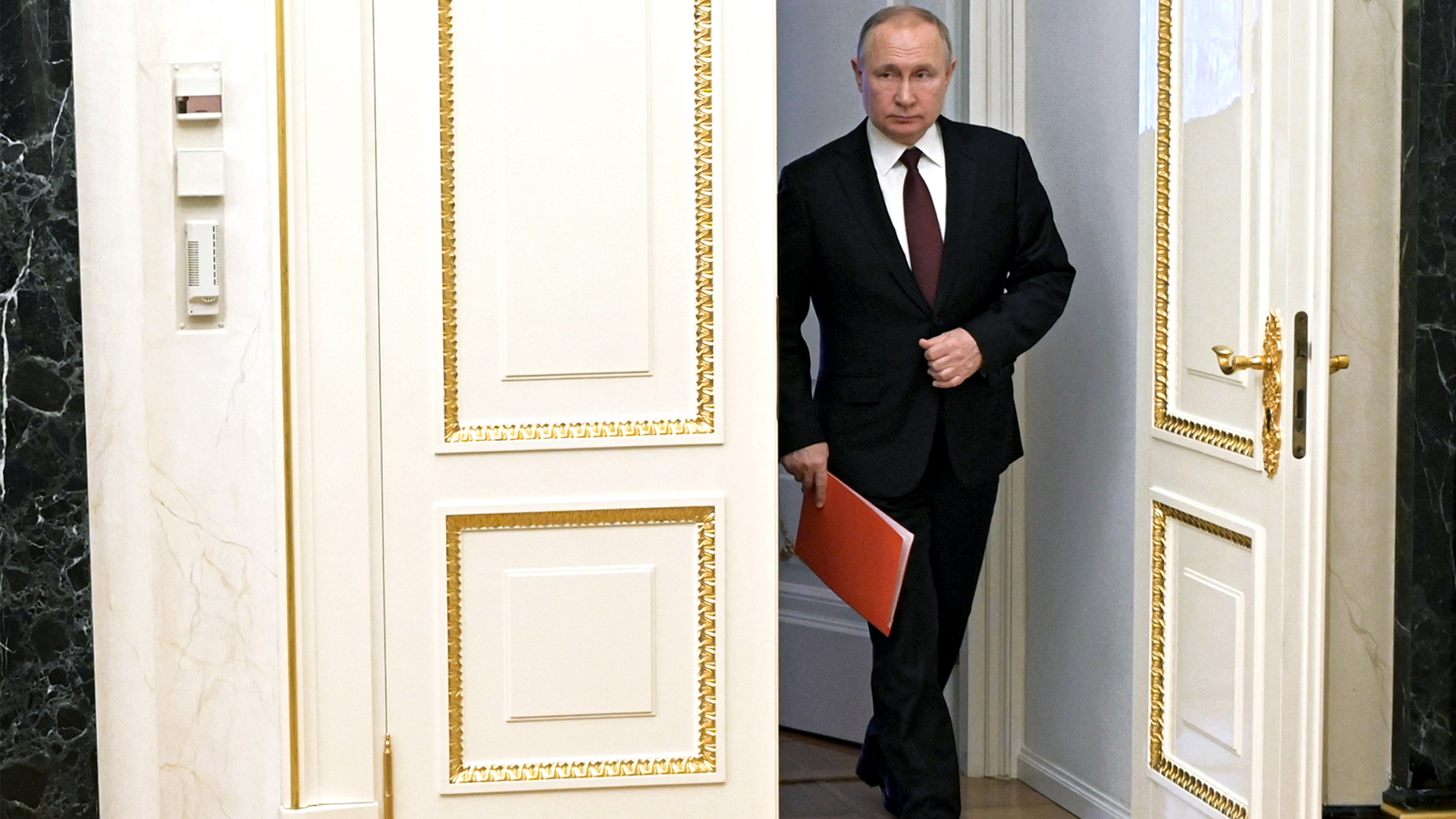 Russian President Vladimir Putin enters a room. He is dressed in a dark suit and tie, and has a red folder in one hand.