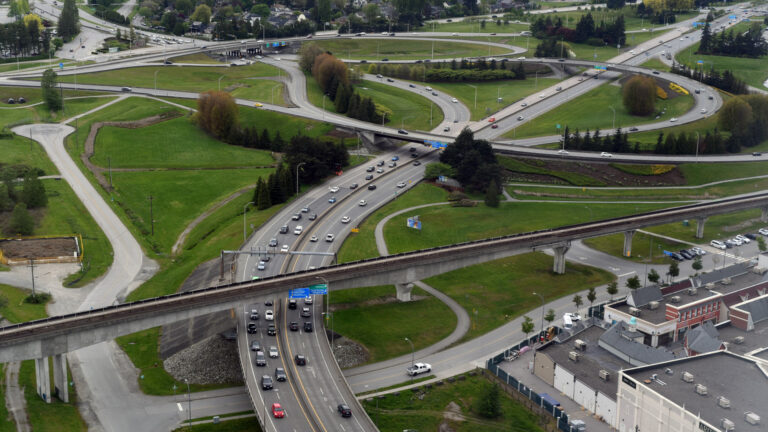 The highway curves to the right and is part of a network of curving highways surrounded by green grass, pockets of evergreens and low-rise buildings here and there.