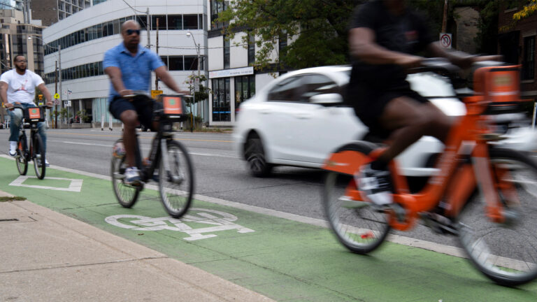 Three cyclists use the lane, which is painted in fading green. A white car passes them.