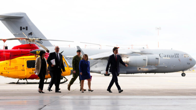 The five political and military leaders walk on the CFB Trenton Tarmac with a yellow-and-red search and rescue helicopter and a grey military transport jet in the background.