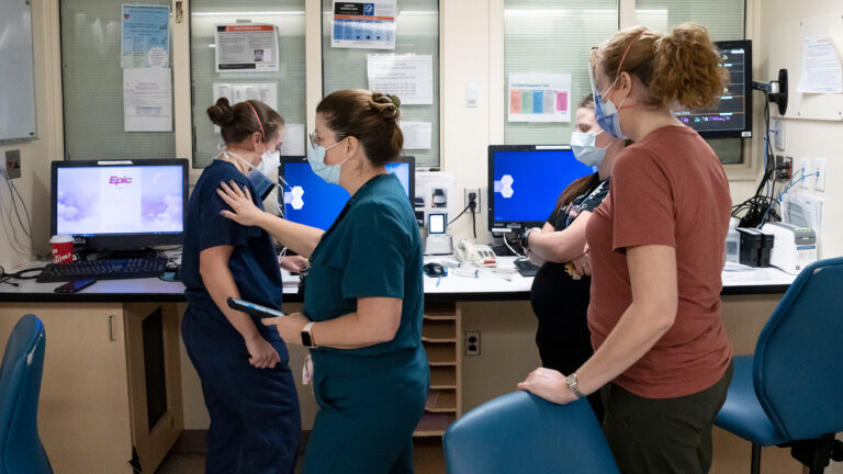 Four women talk in a nurses’ station, where four computers are set up, and paper notices are taped to the walls and windows, which are covered with blinds on the other side of the glass. The women are all masked. One looks at a computer; another has her hand on the first woman’s shoulder.