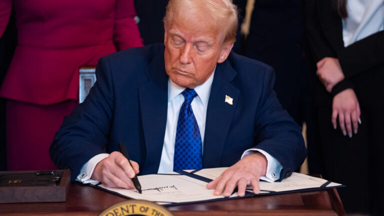 Trump is sitting at a wood desk, leaning forward as he uses a large black marker to make his signature on a two-page document in a black folder. Several people stand behind the desk. He is in a blue suit and tie with a white shirt.