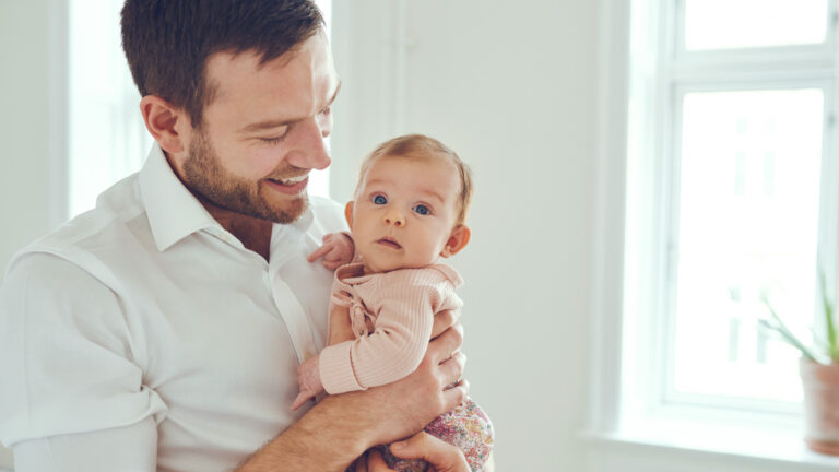 Un homme tient un poupon dans ses bras et le regarde en souriant. L’enfant regarde la caméra.