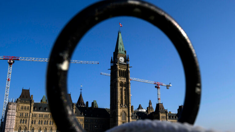 Centre Block and the Peace Tower and construction cranes set against a deep blue sky are seen through a metal fence along the lawn of Parliament Hill.