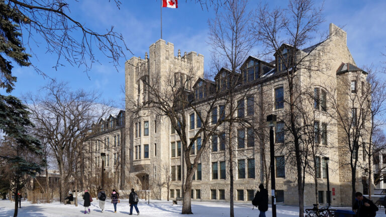 Students walk on the University of Manitoba campus in winter in front of a four-storey limestone clad building.