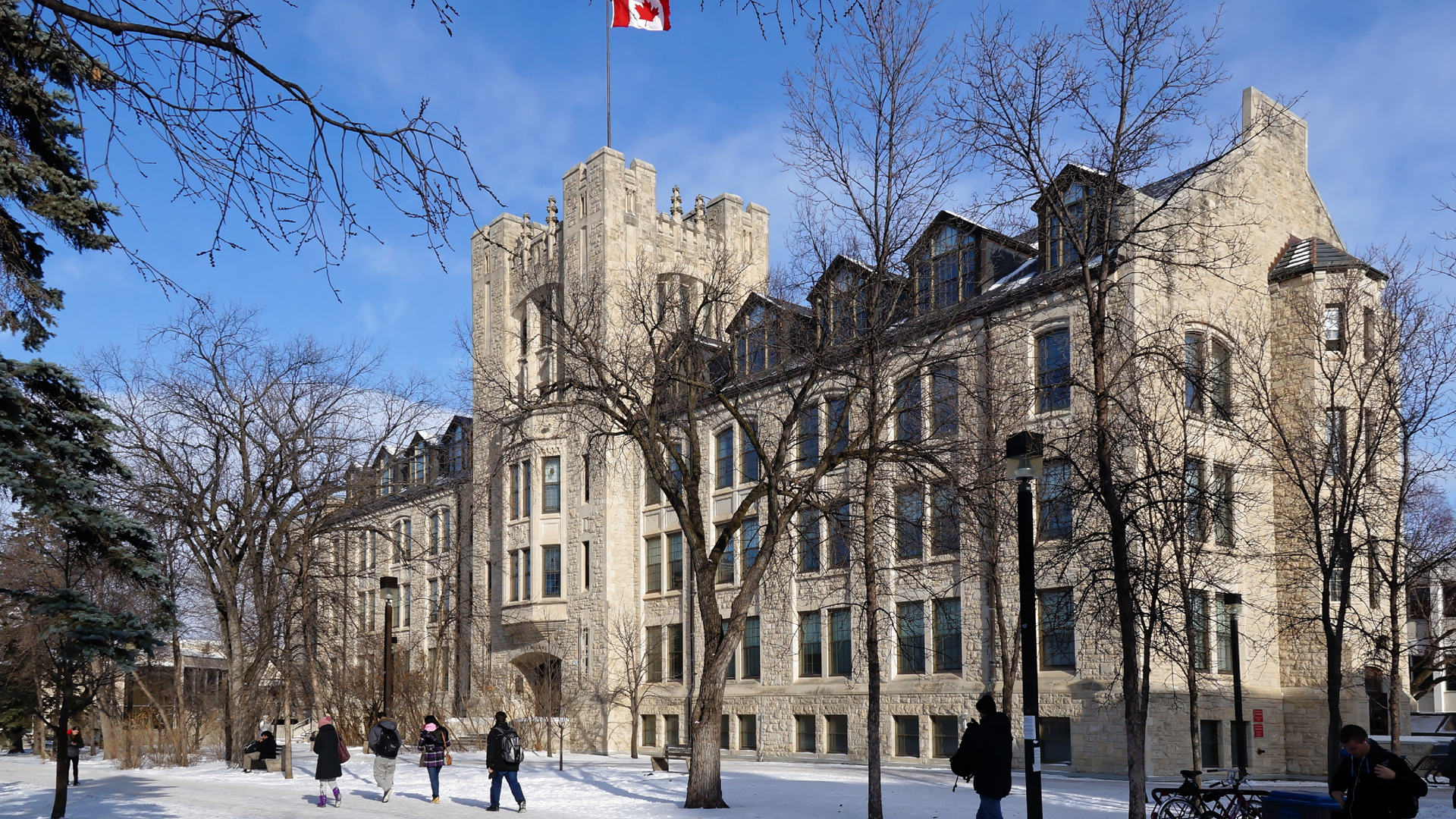 Students walk on the University of Manitoba campus in winter in front of a four-storey limestone clad building.