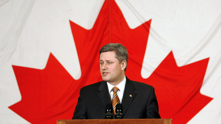 Stephen Harper speaking at a podium in front of a large Maple Leaf.
