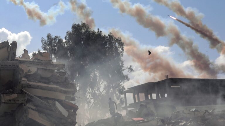 A rocket and several contrails are seen against a blue sky with destroyed concrete buildings below them.