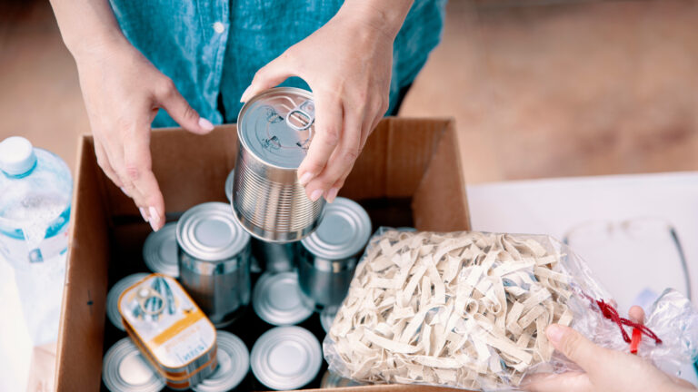 Seen from above sits a box of canned foods. We see the arms and hands of a man holding one of the tins. Another person’s hand holds a clear plastic bag of flat noodles.