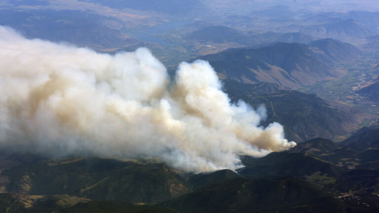 A plume of dense white smoke risks from dark green and brown hills and blows across the frame from right to left. Two valleys with bodies of water and buildings are visible in the distance.