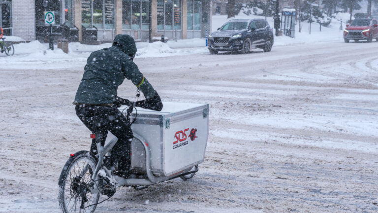 Pushing through snow accumulating on a major downtown street, a man pedals a converted bike whose front end has been converted to carry a large, heavy container for goods.