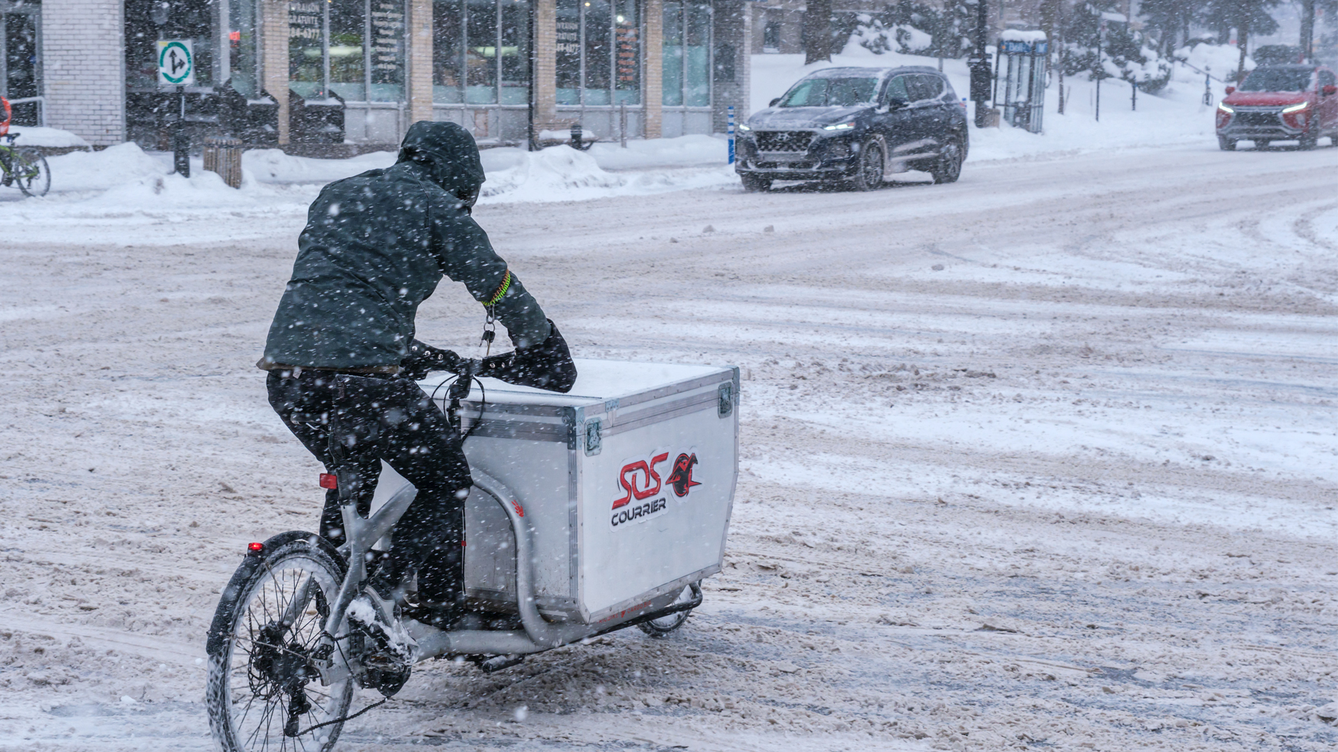 Pushing through snow accumulating on a major downtown street, a man pedals a converted bike whose front end has been converted to carry a large, heavy container for goods.