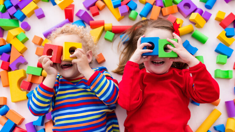Two small children laying among building blocks and looking through blocks with holes in them.