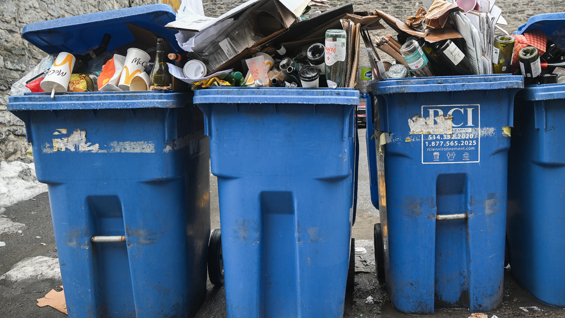 Trois bacs de recyclages bleus sont côte à côte et débordent de contenants en carton et en verre.