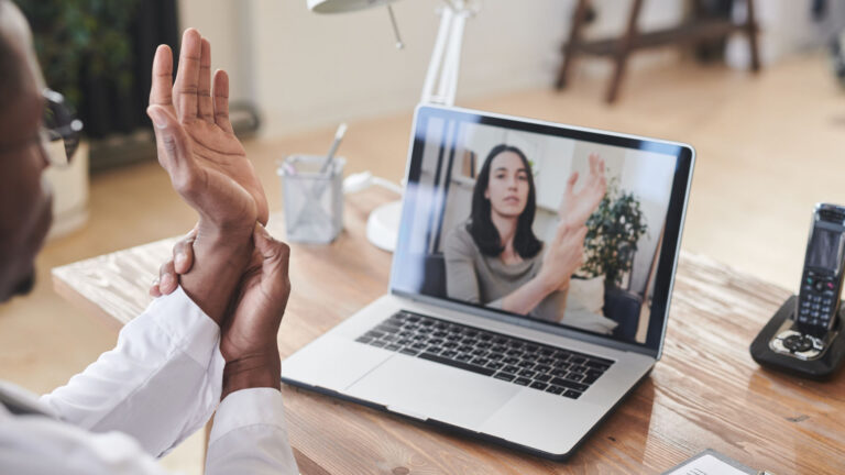 A Black physician at a desk in front of a laptop demonstrates how to apply pressure to the base of his palm in a session with a white woman receiving via the computer.