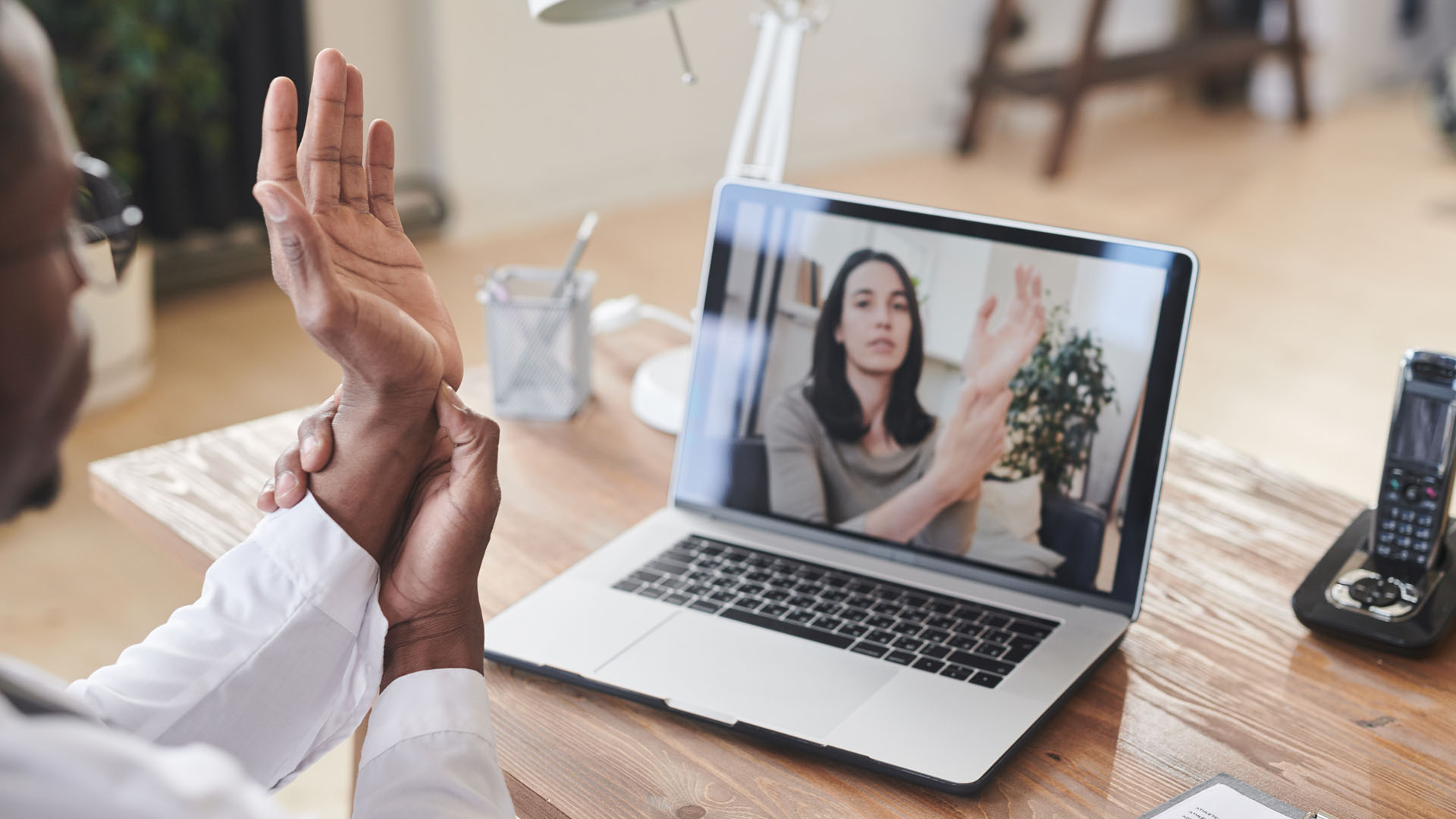 A Black physician at a desk in front of a laptop demonstrates how to apply pressure to the base of his palm in a session with a white woman receiving via the computer.