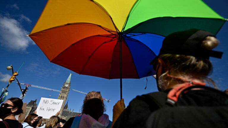 A protester holds up an umbrella in the colours of the rainbow against a blue sky with the Peace Tower of Parliament in the background.
