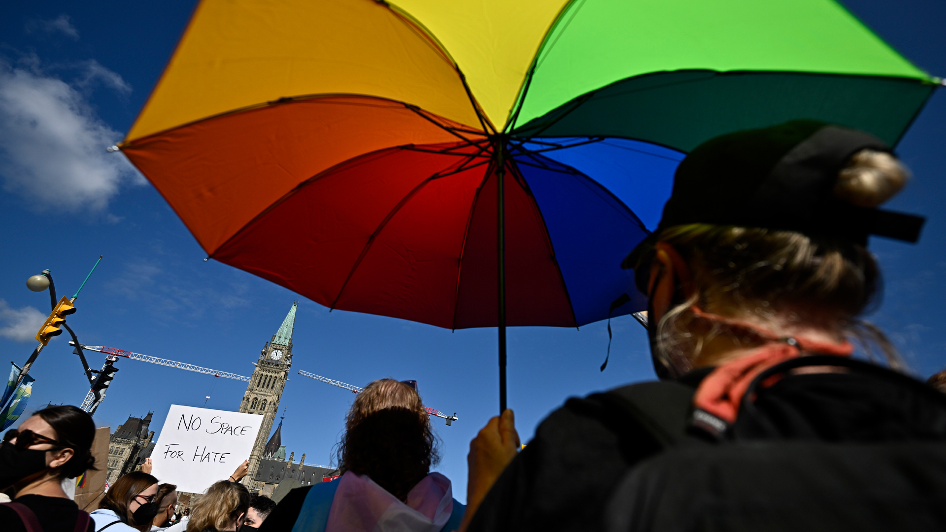 A protester holds up an umbrella in the colours of the rainbow against a blue sky with the Peace Tower of Parliament in the background.