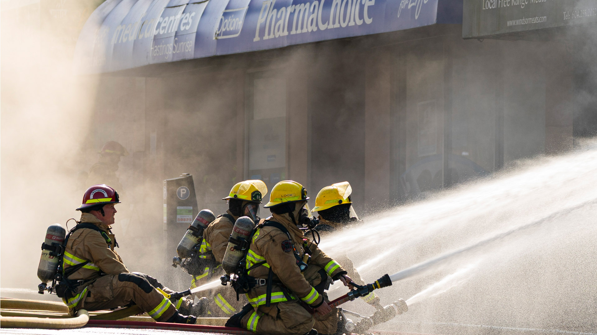 Four firefighters sit and kneel on hoses spraying water on a building surrounded by smoke and mist.